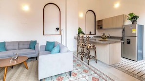 Een moderne woonkamer met een grijze bank, een witte fauteuil en een kleine houten salontafel. In de achtergrond is een keuken zichtbaar met een koelkast en een spoelbak.