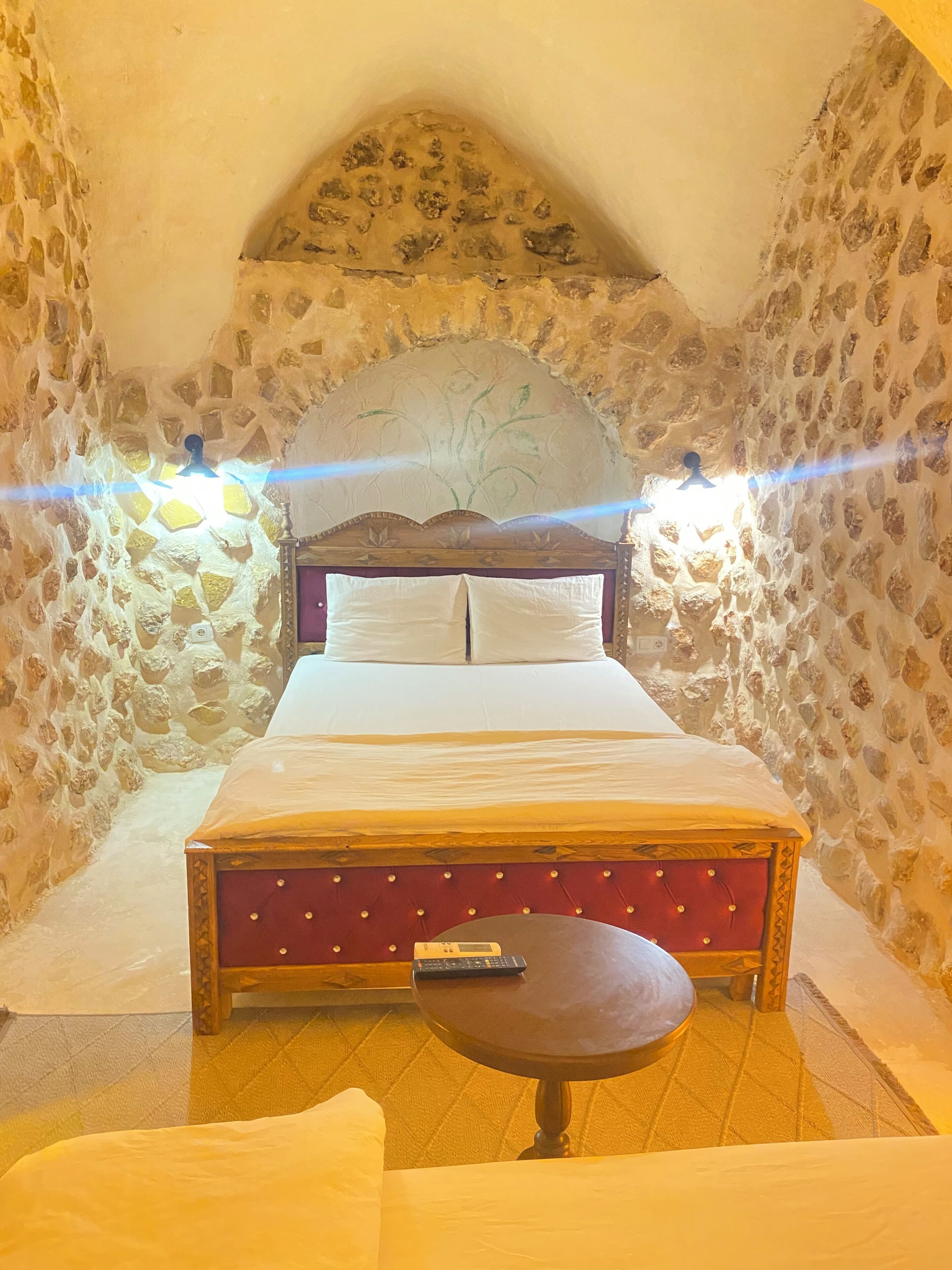 A bedroom with a stone wall, a wooden bed, a small round table, and a ceiling light.