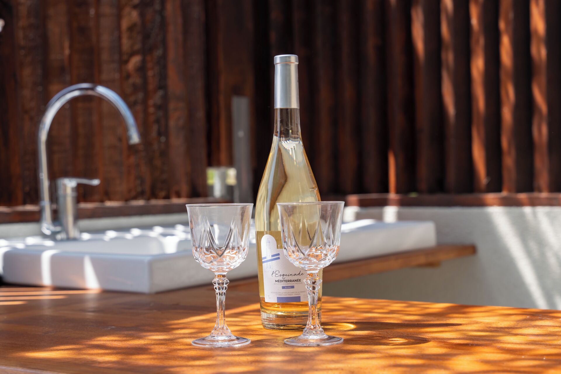 A bottle of wine and two glasses on a wooden table with a sink and faucet in the background.