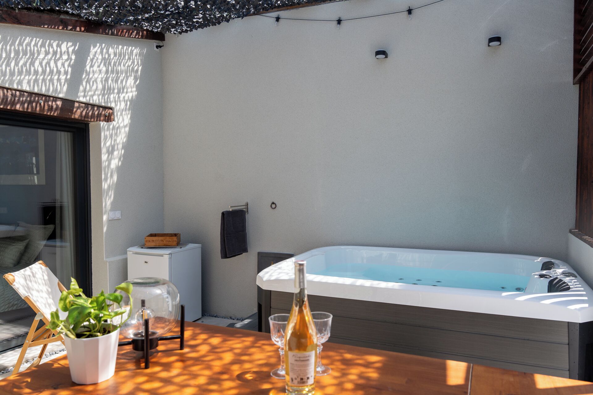Une terrasse ensoleillée avec un jacuzzi, une table avec des verres et une bouteille, et une plante en pot.