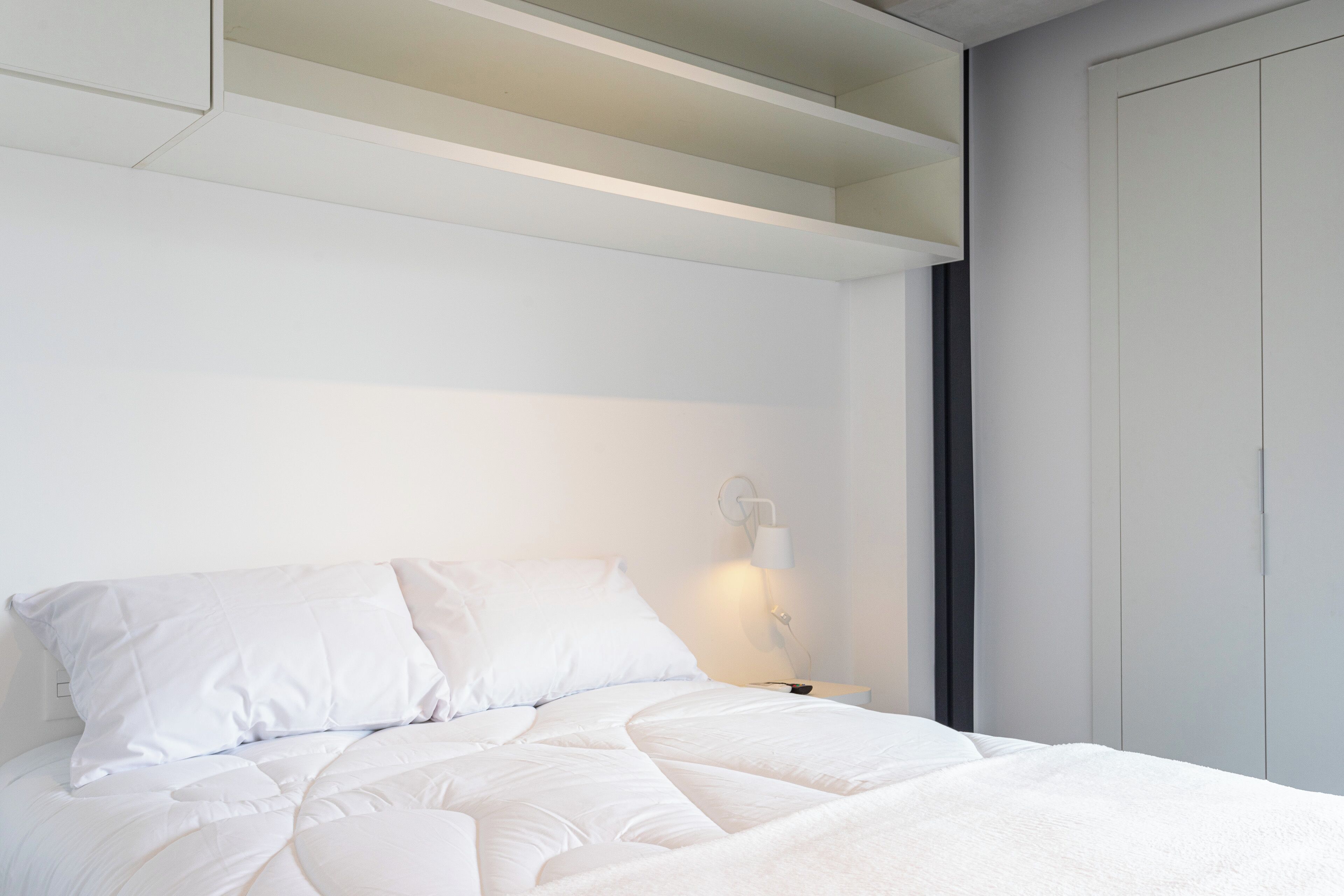 Un llit ben arriscat amb roba de llit blanca en una habitació moderna, amb un armari encastrat i una tauleta de nit amb una làmpada.