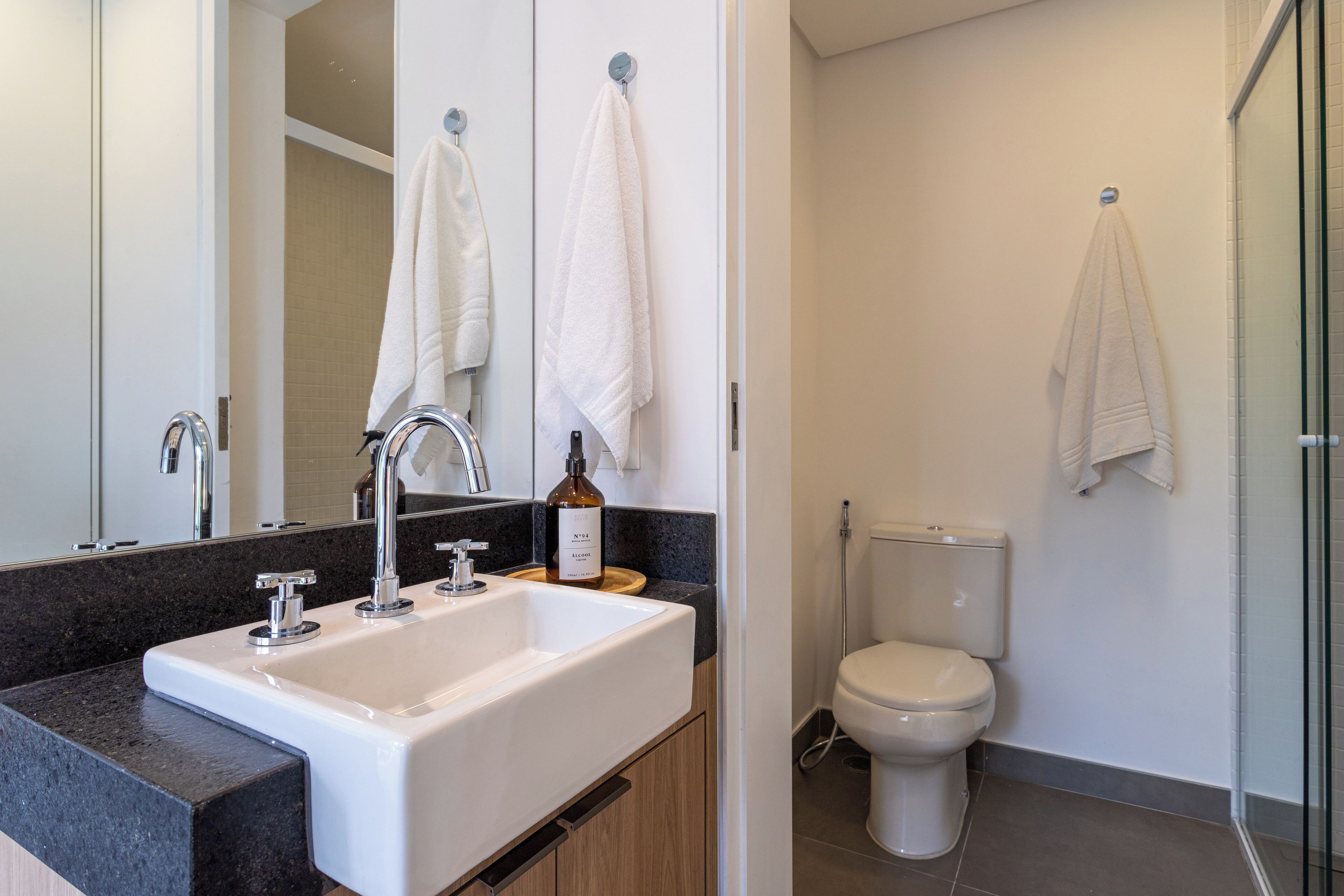 A modern bathroom with a white sink, two faucets, a bottle of hand soap, and a toilet.