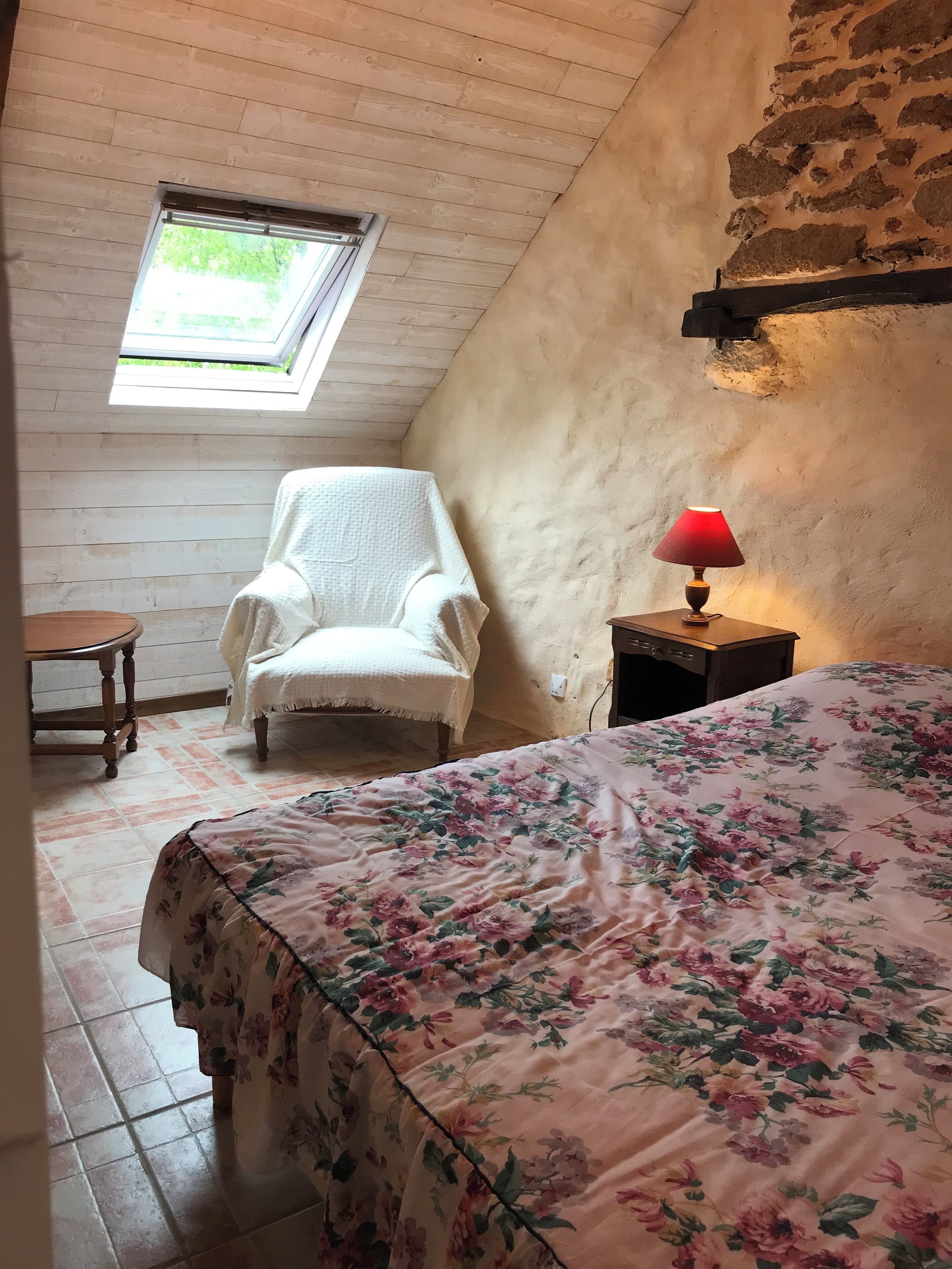 A cozy room with a sloped ceiling, a wooden chair, a bed with a floral bedspread, a small table, and a lamp.