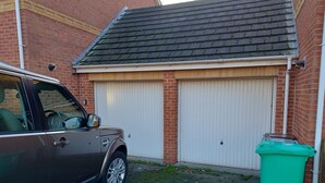 Un garaje con dos puertas blancas, un edificio de ladrillo y un coche aparcado.