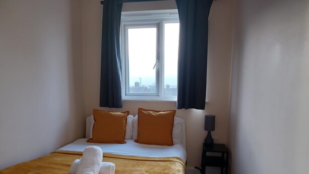 Habitación de hotel con cama, dos almohadas naranjas, una manta blanca, una toalla blanca, una ventana con cortinas y una pequeña mesa con lámpara.