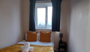 A hotel room with a bed, two orange pillows, a white blanket, a white towel, a window with curtains, and a small table with a lamp. - Marvyn-Five House- Huku Kwetu (Nottingham)
