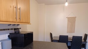 Una cocina con armarios de madera, un microondas negro, un cesto de basura blanco y una mesa de comedor con sillas negras.