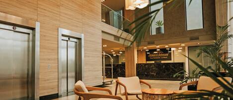 A lobby with wooden panel walls, two elevators, a reception desk, and a seating area with wicker furniture.