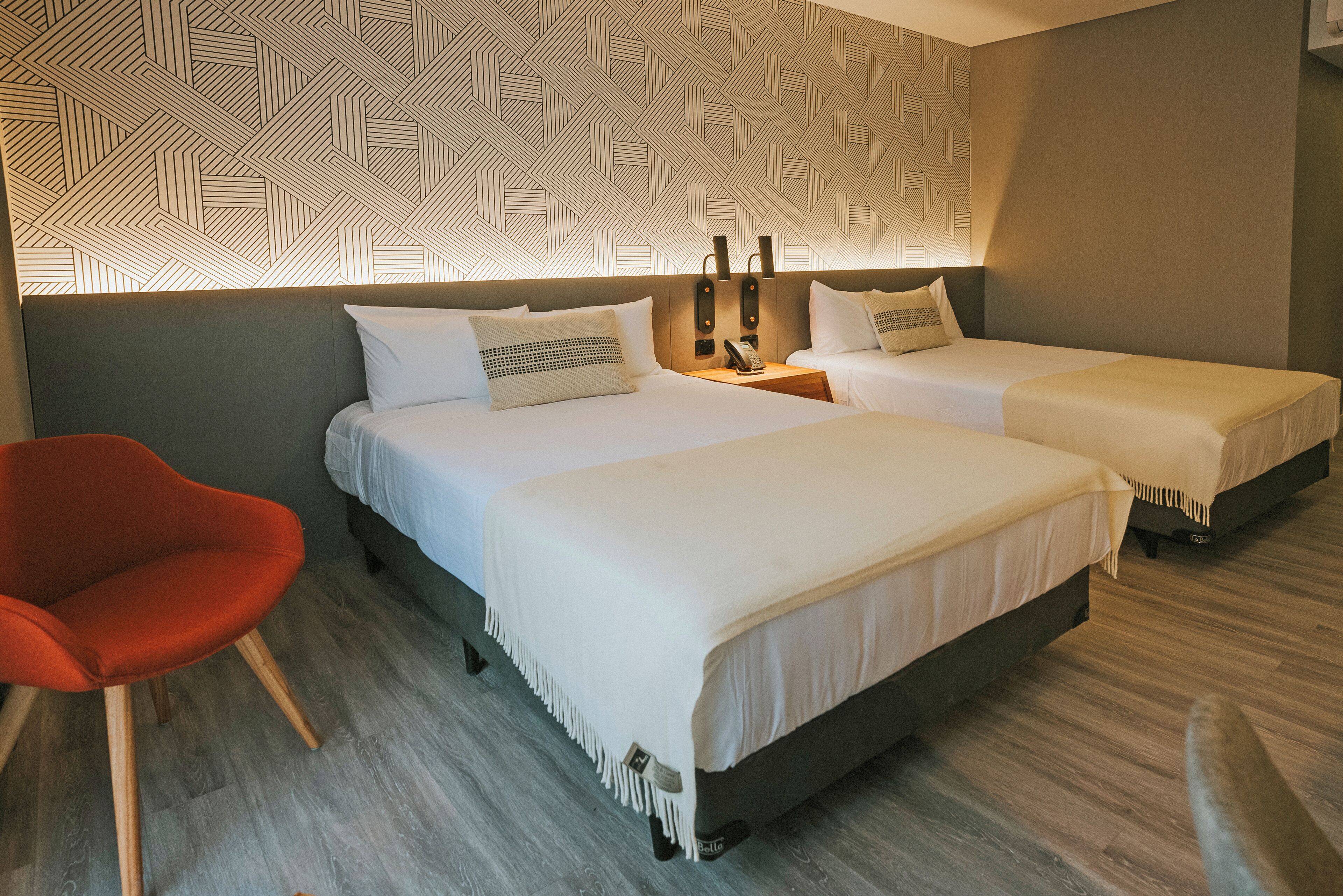 A hotel room with two beds, a red chair, and a patterned wall.