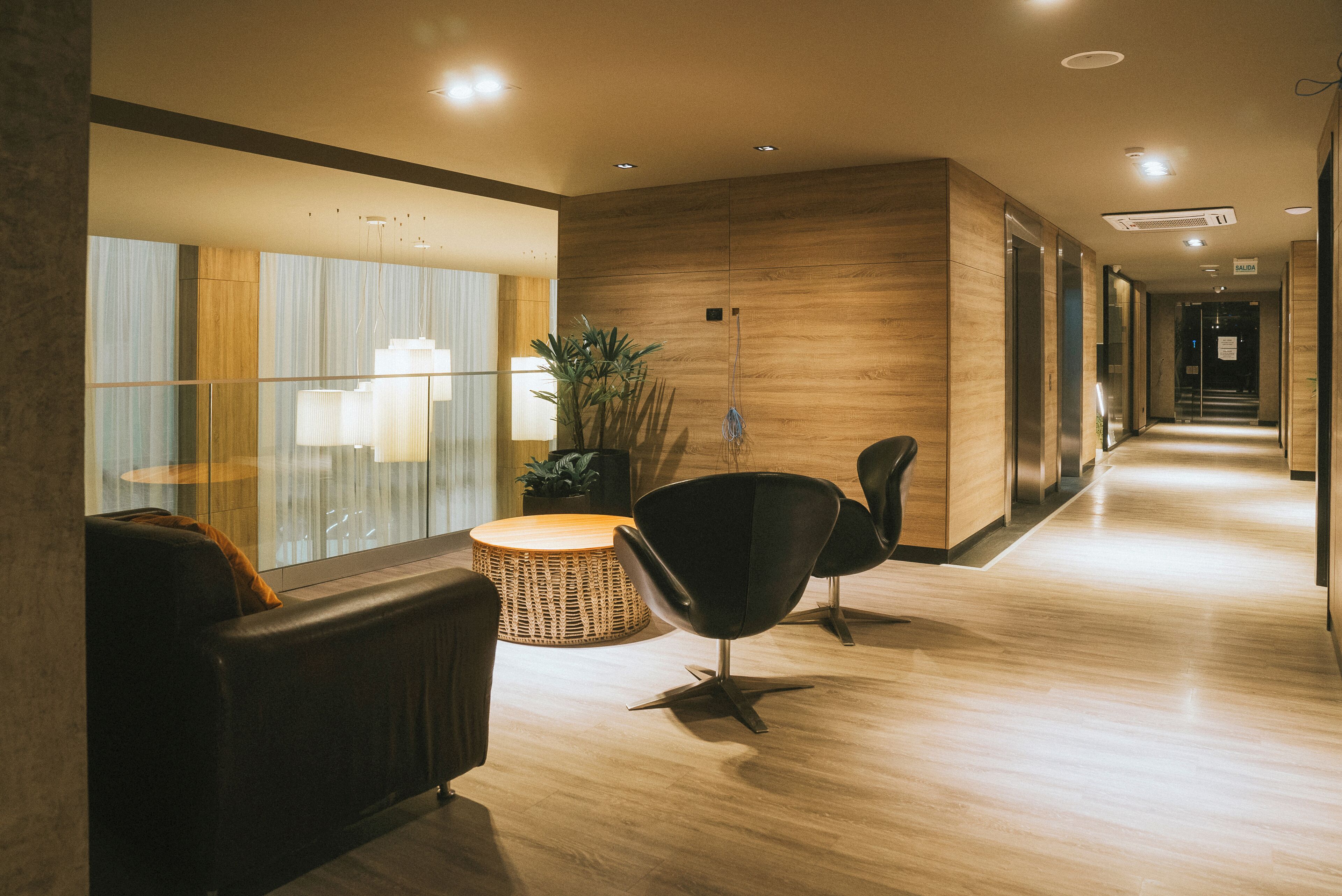 A modern hotel lobby with a wooden floor, leather seating, a wicker table, and a large window with curtains.