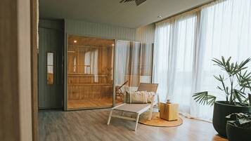 A modern room with a wooden floor, a glass sliding door leading to a balcony, a white chair with cushions, and large potted plants.