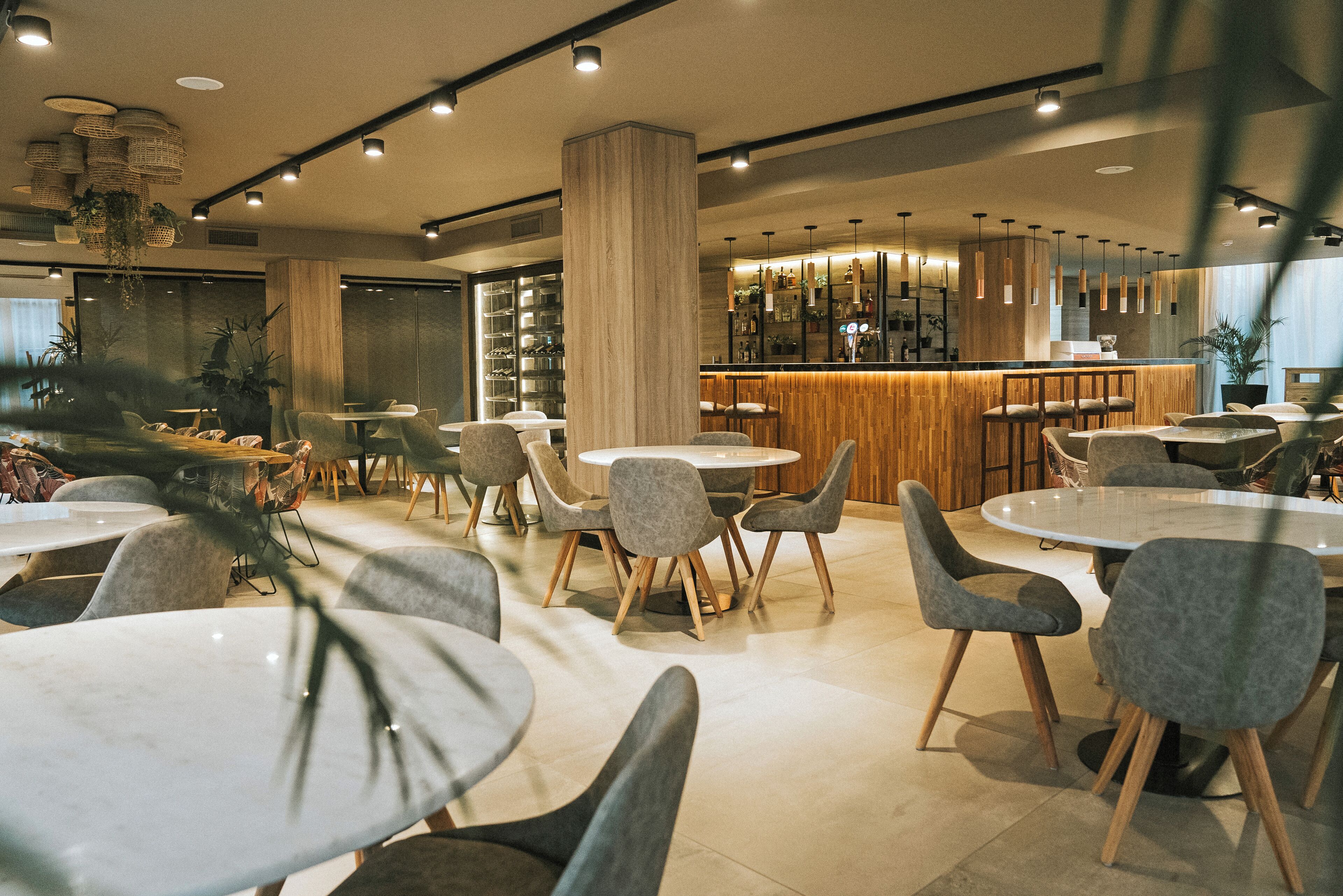 Un restaurante moderno con mesas redondas y sillas grises, una zona de bar de madera y lámparas colgantes.