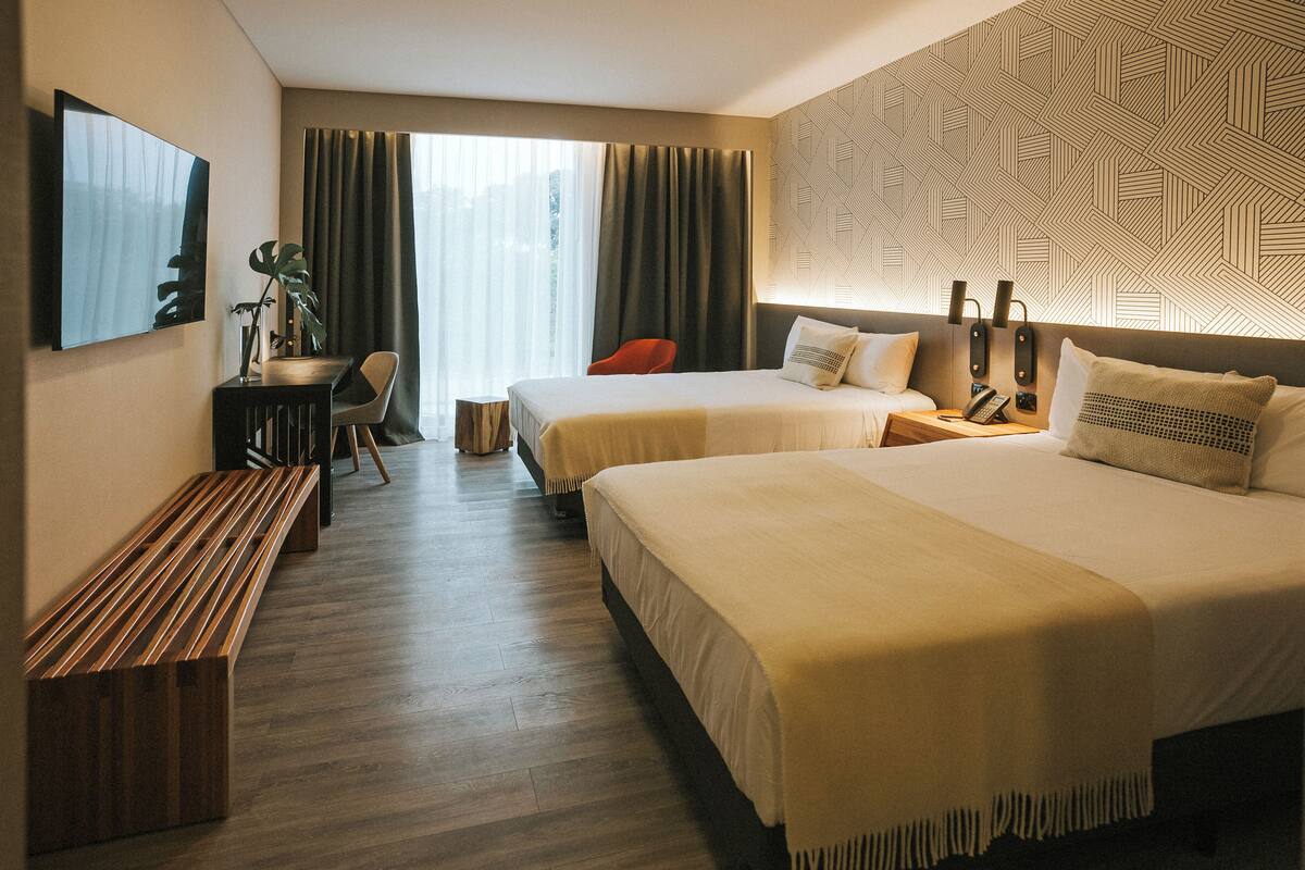 A hotel room with two beds, a desk, a chair, and a bench.