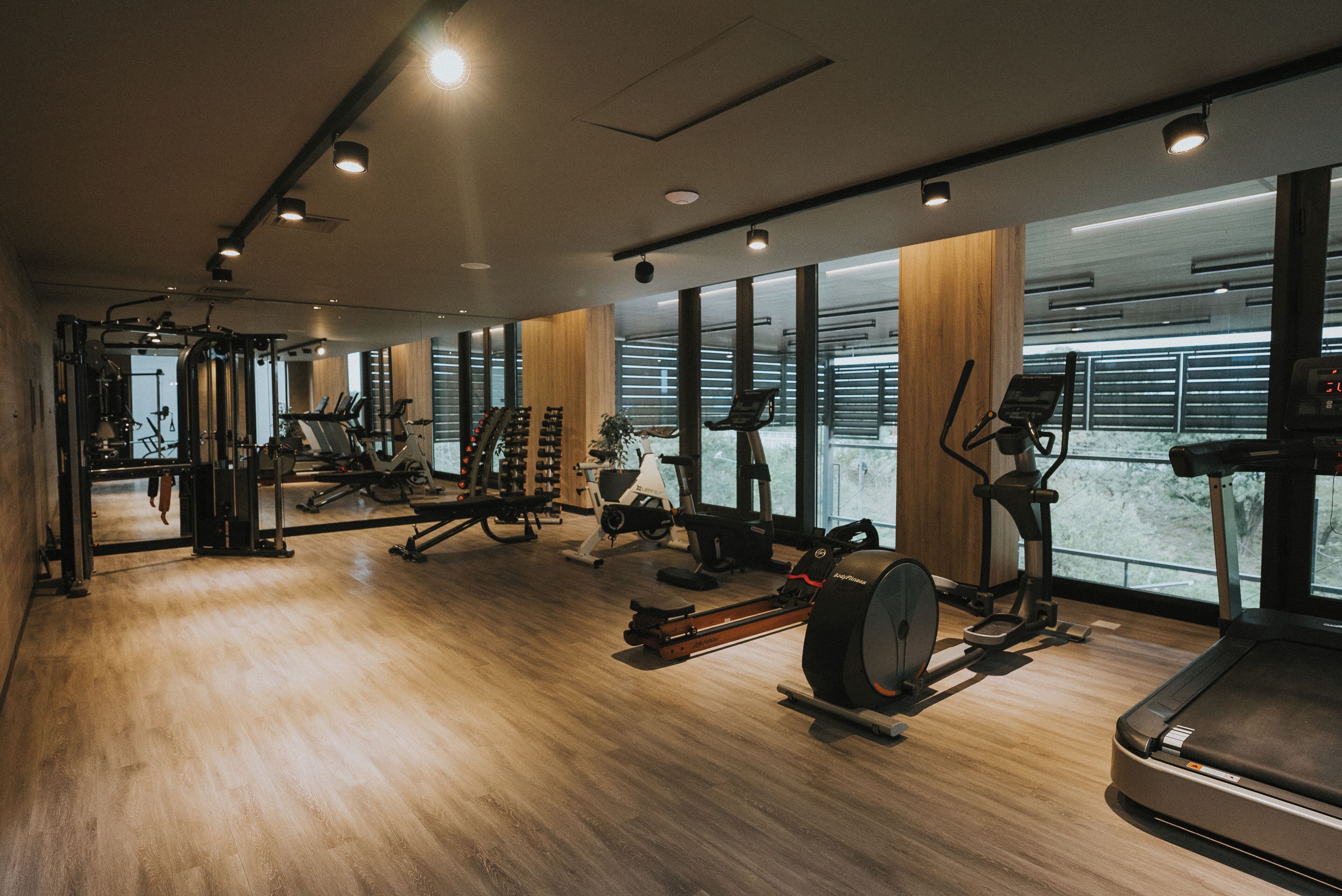 Un gimnasio moderno con diversos equipos de ejercicio, amplias ventanas y suelo de madera.