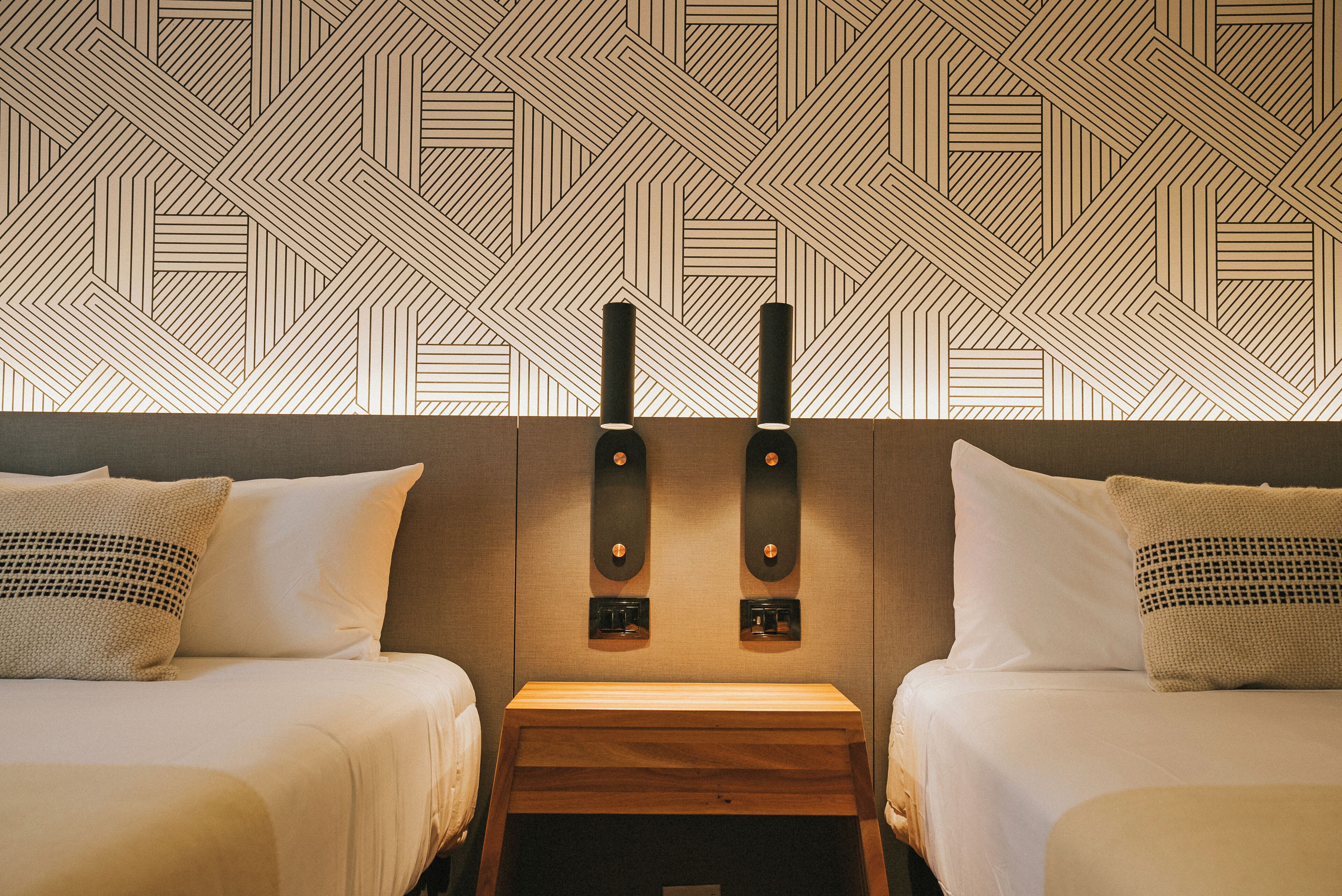 Habitación de hotel con dos camas, una mesita de noche de madera y un papel tapiz estampado.