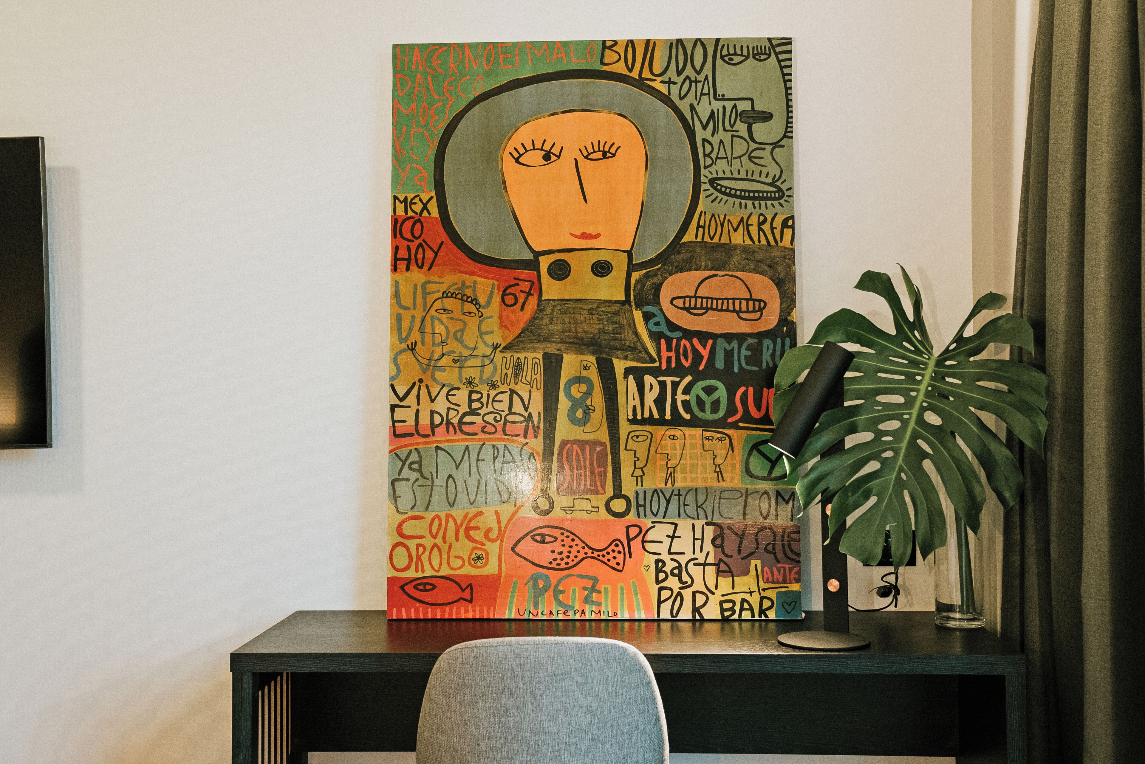 Un escritorio con un cuadro abstracto colorido, una planta en maceta y una silla.