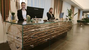 Ein Empfangsschalter mit Glastischplatte und Holzfuß, zwei Personen dahinter sowie ein YASHI-markierter Computermonitor.