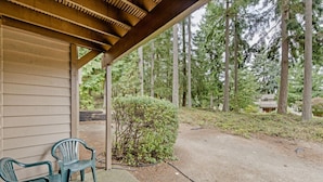 Uma área externa coberta com duas cadeiras de plástico verde, uma pérgola de madeira e uma trilha de terra que leva a uma área arborizada.