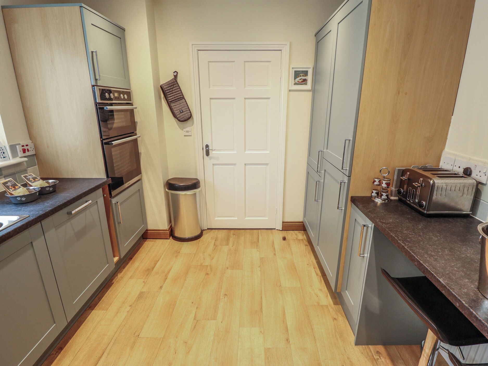 A compact kitchen with a stove, oven, and toaster. There is a trash bin and a door with a small picture on the wall.