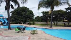 A swimming pool with a slide, surrounded by palm trees and a few chairs. - Moba Hotel & Convention Centre (Kitwe)