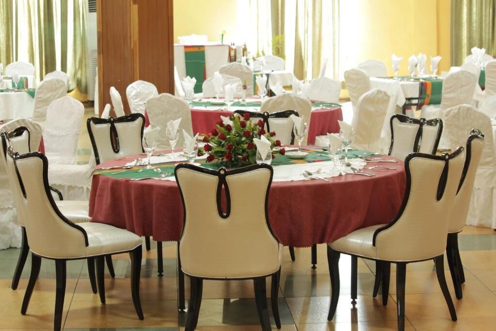 A banquet hall with round tables covered in red tablecloths, surrounded by white chairs with black trim.