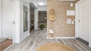 A hallway with a wooden floor, white walls, and a wooden accent wall. There are two doors on the left, a mirror on the left wall, and a hat rack with hats and a handbag on the right wall. - Upland Home I by Madeira Sun Travel (Ponta do Sol)