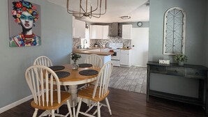 A dining area with a wooden table and white chairs, a chandelier, and a kitchen in the background. - Cheerful Bronson Cutie
 (Bronson)
