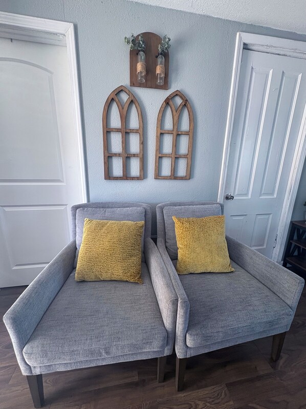 Deux fauteuils gris avec des coussins jaunes, une décoration murale en bois avec deux fenêtres en arc de cercle, et une porte blanche.