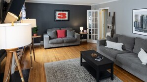 A modern living room with a grey sofa, a black coffee table, and a grey rug. There is a framed number '3' on the wall, a lamp, and a door leading outside. - Charming Old Orchard home with fenced yard (Toledo)