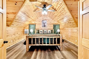 Une chambre dans un chalet en rondins, avec un lit placé au centre, sous un plafond en pente, un ventilateur de plafond et un tableau encadré au mur.
