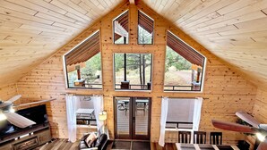A cozy cabin interior with a wooden interior, a fireplace, a dining area, and large windows offering views of the surrounding forest. - Modern & Private Cabin Retreat Near Sitgreaves National Forest! (Overgaard)