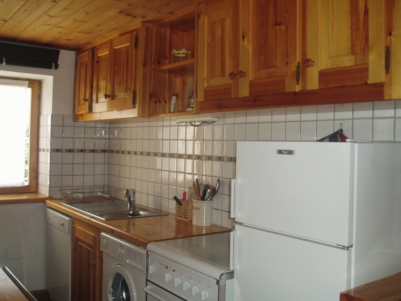 Une cuisine avec des meubles de rangement en bois, un pare-feu en carrelage, un évier, une cuisinière, un lave-linge et un réfrigérateur.
