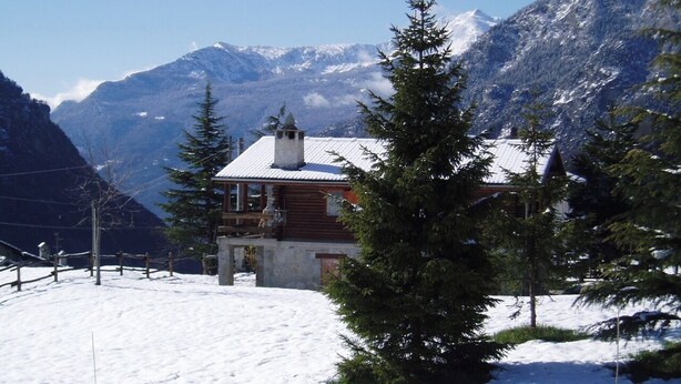 A wooden cabin with a snow-covered roof, surrounded by snow and mountains.
