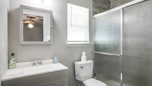 A bathroom with a white sink, mirror, and toilet, a window with blinds, and a shower area with frosted glass doors. - 4 bed/ 2 bath gem
 (Killeen)