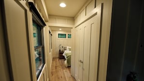 Een smalle gang met houten vloer, witte wanden en deuren die leiden naar een slaapkamer met een bed en een raam.