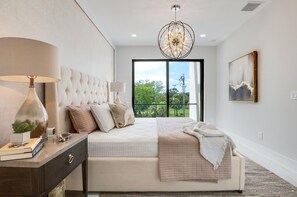 Ein Schlafzimmer mit einem großen Bett, einem Nachttisch, einer Lampe, einem Gemälde und Blick auf Grün durch das Fenster.