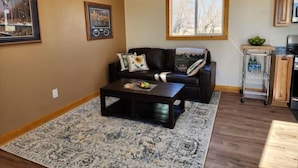 A living room with a dark brown leather sofa, a coffee table, and a patterned rug. There is a window with a view of trees and a kitchen area with a metal cart. - The Bunk House (Cody)