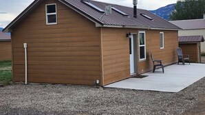 A single-story wooden building with a sloped roof, a small porch with two chairs, and a gravel driveway. - The Bunk House (Cody)