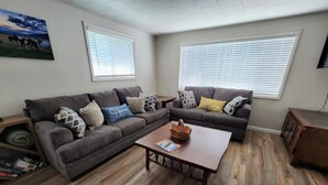 A living room with a sectional sofa, a coffee table, and a wooden floor. There are two windows with blinds, a painting on the wall, and a small wooden cabinet. - Easy Living ~ Easy Playing, just a 5 minute walk from down town Cody. (Cody)