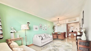 A living room with a green wall, a white sofa, a dining area with a table and chairs, and a kitchen in the background.