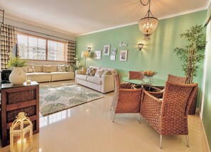 A living room with a sofa, a dining table with chairs, a chandelier, and a large window with blinds.