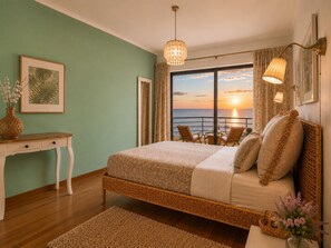 A bedroom with a large bed, a desk, a chair, a view of the sunset over the sea, and a hanging light fixture.