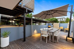 Een terras buiten met een houten dek, een tafel met stoelen, een grote parasol en een barbecue.