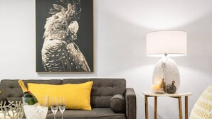 A living room with a grey sofa, a yellow pillow, a table with a bottle of champagne, glasses, and a fruit platter.