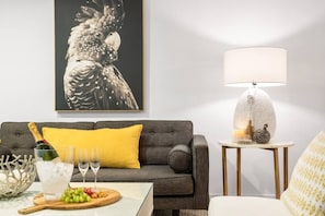 Una sala de estar con un sofá gris, una almohada amarilla, una mesa con una botella de champán, copas y una bandeja de frutas.