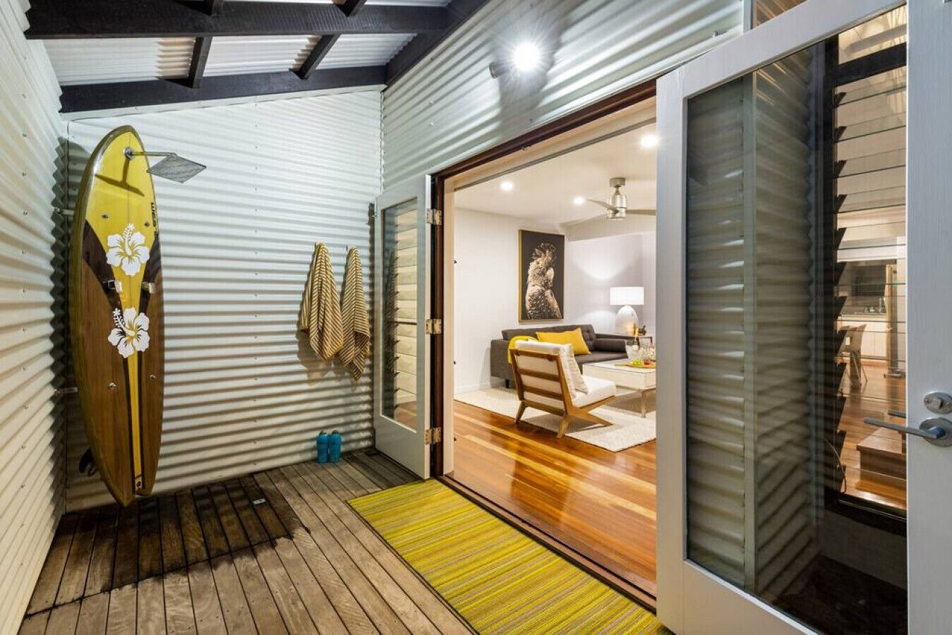 A compact living space with a wooden floor, a surfboard leaning against a corrugated wall, and a sliding glass door leading to a bedroom with a bed and a painting.