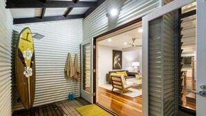 A compact living space with a wooden floor, a surfboard leaning against a corrugated wall, and a sliding glass door leading to a bedroom with a bed and a painting.
