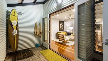 A compact living space with a wooden floor, a surfboard leaning against a corrugated wall, and a sliding glass door leading to a bedroom with a bed and a painting.