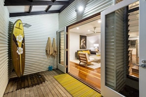 Un espacio habitable pequeño con suelo de madera, una tabla de surf apoyada contra una pared de metal corrugado y un ventanal corredizo que da a un dormitorio con cama y un cuadro.