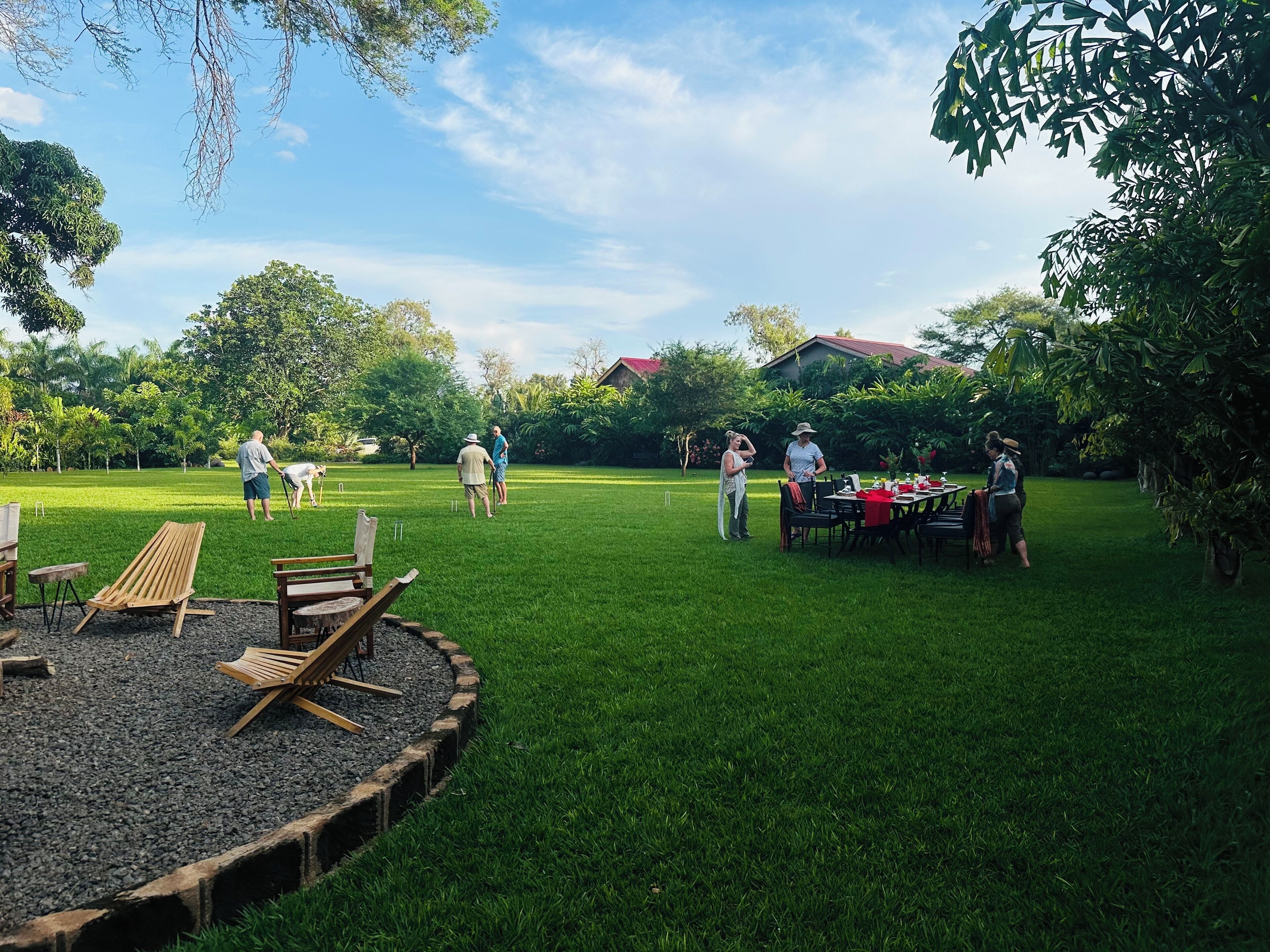 A grassy outdoor area with a table set up for dining, surrounded by trees and a house in the background.